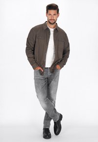 Donker olijf groene knoopjack met twee borstzakken, een witte t-shirt eronder, lichtgrijze spijkerbroek en zwarte schoenen. Gladde textuur.