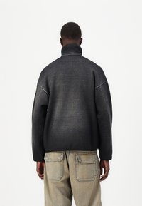 Muž v tmavě šedém vysokém límci a béžových cargo kalhotách, stojící zády k fotoaparátu na jednobarevném pozadí.