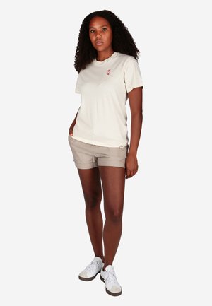 Frau mit lockigem Haar trägt beige Shorts, off-white T-Shirt mit kleinem rotem Blumenmotiv und weißen Sneakers, steht mit einer Hand in der Tasche.