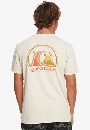 Mies, joka pukee beige T-paidan, jossa on punainen ja keltainen aalto- ja tulivuorigrafiikka sekä "Quiksilver The Original Surf Co." -teksti selässä.