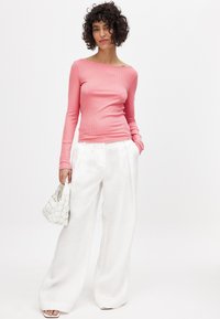 Rozā ribota garām piedurknēm blūze, apvienota ar baltām platām biksēm. Modelis tur nelielu baltu austu somiņu, demonstrējot tīru, moderno izskatu.