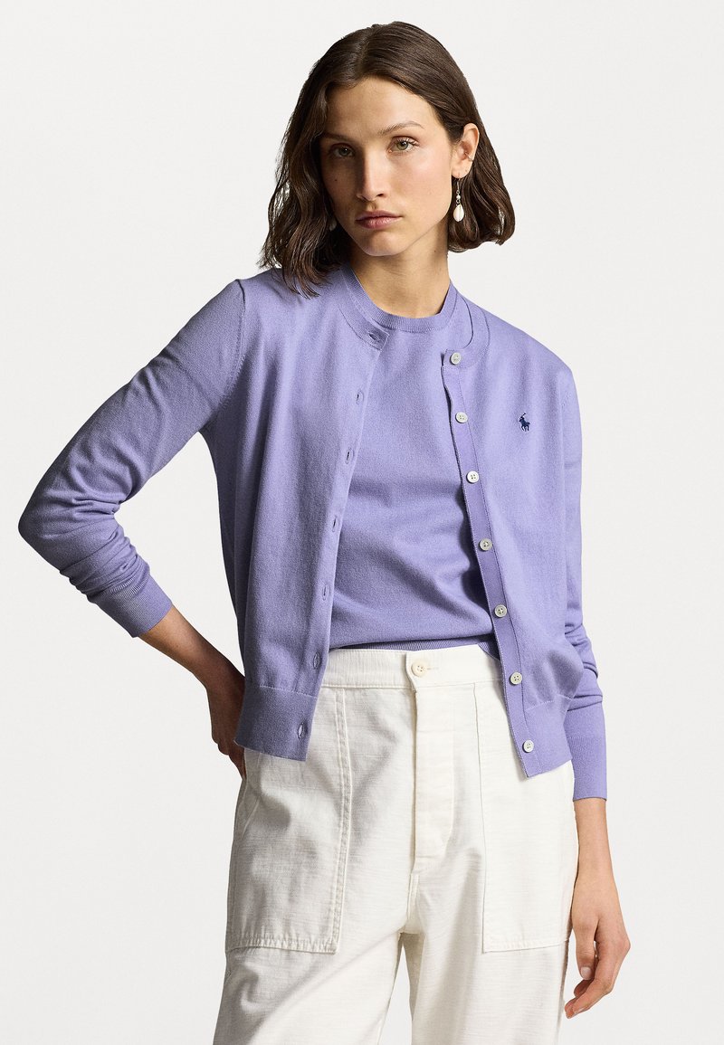 Cardigan violet clair à manches longues et col rond, avec des boutons sur le devant et un petit logo brodé sur la poitrine. Associé à un pantalon crème.