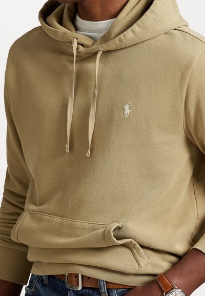 Persoon in een beige hoodie met trektouwen, witte onderhemd, blauwe jeans, een bruine leren riem en een bruine leren polshorloge.