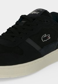 Μαύρο sneaker Lacoste με λευκό λογότυπο στο πλάι, μαύρα κορδόνια, υφασμένο επάνω μέρος και εκρού σόλα σε απλό φόντο.