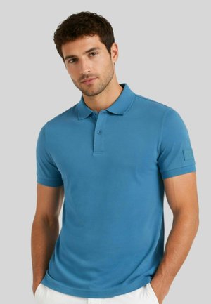 Uomo che indossa una polo blu a maniche corte con colletto e patta con bottoni, in piedi con le mani nelle tasche su sfondo grigio.