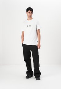 Vit t-shirt med kort ärm och grafiska tryck, tillsammans med svarta vida byxor och svarta slip-on skor. Minimalistisk design, slät textur.