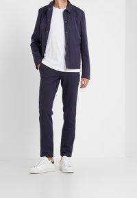 Marineblå letvægtsjakke med trykknapper, båret over en hvid T-shirt og parret med matchende marinebukser og hvide sneakers. Glat tekstur.