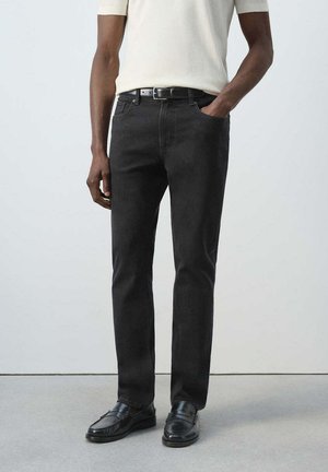 Man wearing straight black jeans, black belt, black loafers, and a short-sleeve cream shirt with one hand in pocket.