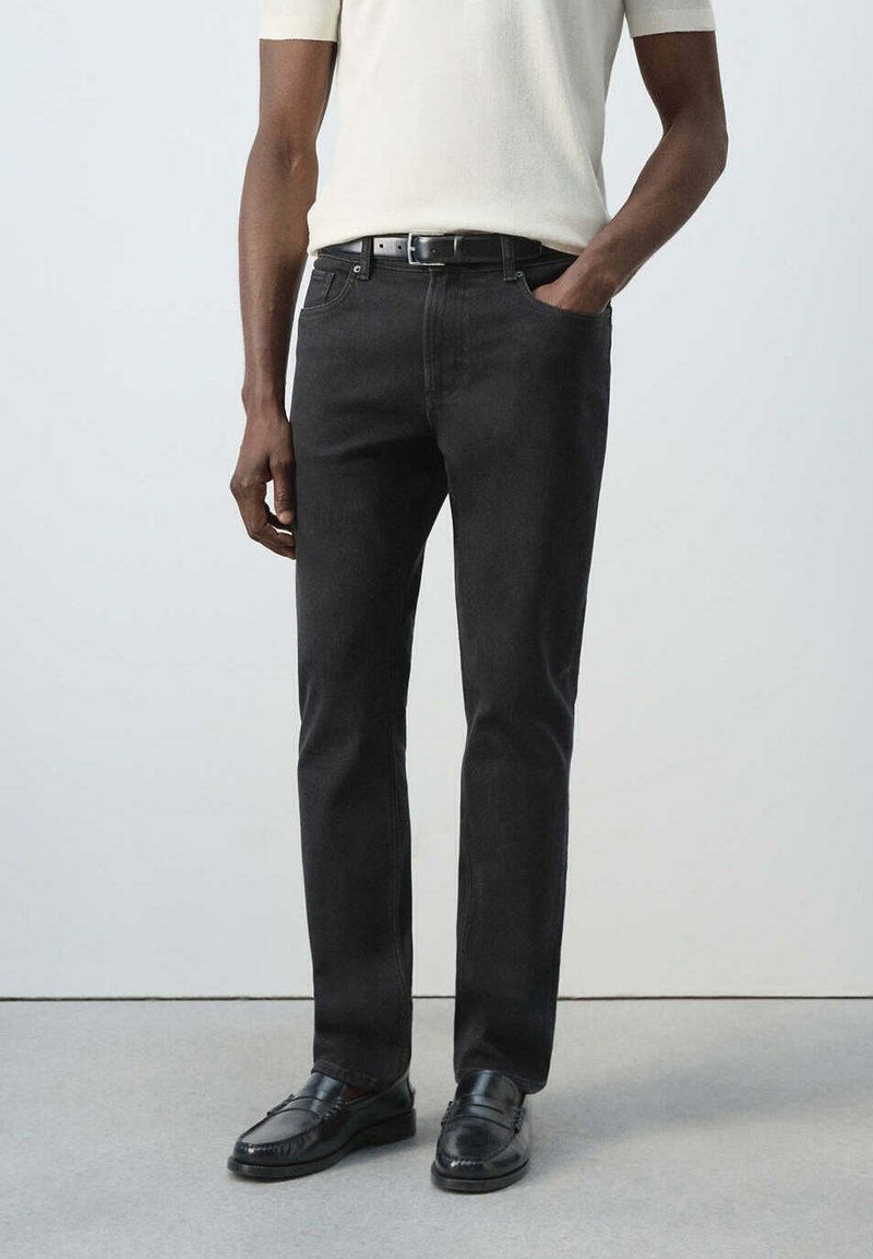 Homme portant un jean noir droit, une ceinture noire, des mocassins noirs et une chemise crème à manches courtes, une main dans la poche.
