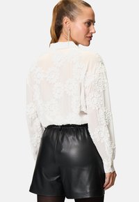 Blouse blanche avec des détails en texture florale, dotée de manches longues et d'un col. Associée à un short en cuir noir taille haute.
