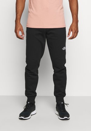 Mann trägt schwarze The North Face Jogginghose, schwarze Adidas Sneakers und ein hellrosa T-Shirt, steht vor weißem Hintergrund.