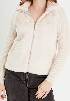 Femme portant une veste crème zippée avec un devant en polaire douce et des manches tricotées, associée à un jean gris foncé, debout, main droite près de la hanche.