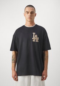 Camiseta de algodón azul marino oscuro con un gran logo texturizado "LA" en el pecho. La camiseta tiene un corte relajado y un cuello redondo acanalado.