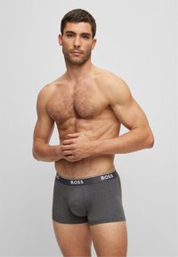Graue Boxershorts mit einem schwarzen Logo-Bund, ausgestattet mit einem glatten, dehnbaren Stoff und einem figurbetonten Design, präsentiert auf einem nackten Oberkörper.