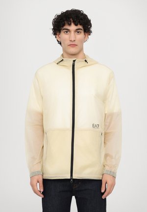 Lightweight cream windbreaker jacket with a hood, black zip closure, elastic cuffs, and EA7 logo on the front. Smooth texture, slightly transparent.