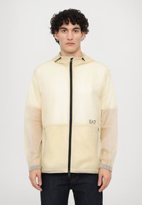 Lightweight, cream-colored jacket with a hood, black zipper, and logo accent. Features elastic cuffs and a smooth, translucent texture.