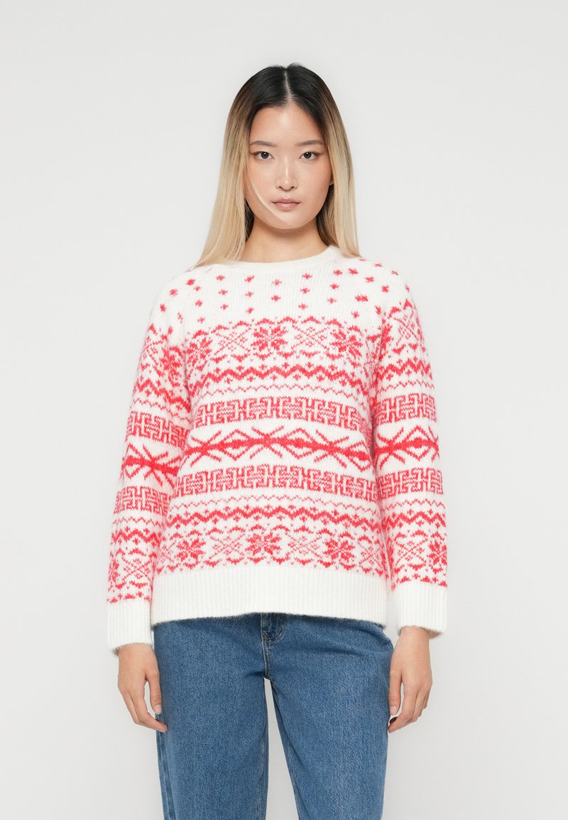 Mulher com cabelo liso a usar um suéter branco com padrões nórdicos vermelhos e jeans azuis, de pé contra um fundo branco simples.