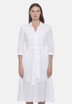 Donna che indossa un vestito bianco al ginocchio con bottoni, motivo floreale a occhiello, maniche a tre quarti, colletto e cintura annodata in vita.