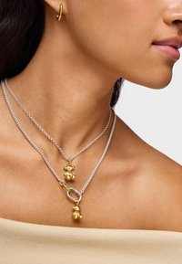 Złote i srebrne naszyjniki warstwowe z zawieszkami w kształcie niedźwiedzia i detalem ogniw, o delikatnej teksturze łańcuszka i gładkim metalicznym wykończeniu.