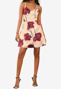 Bloemenjurk met een V-hals en dunne bandjes. Kenmerkt zich door rode bloemen op een beige achtergrond, elastische taille en een uitlopende rok.