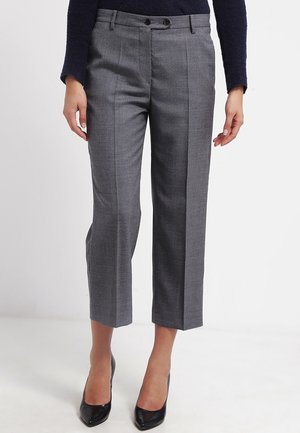 Frau trägt graue maßgeschneiderte, knöchellange Hose mit Bügelfalten vorne und schwarzen High Heels, steht vor einem schlichten Hintergrund.