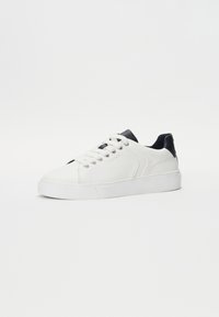 Adidași din piele albă cu accente negre în călcâi. Au vârf rotund, design cu șireturi și o talpă din cauciuc texturată. Logo-ul mărcii este vizibil pe lateral.