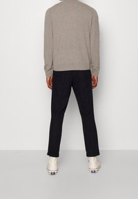 Osoba stojąca przodem do ściany, ubrana w beżowy sweter, czarne spodnie z mankietami oraz białe sneakersy wysokie z logo.