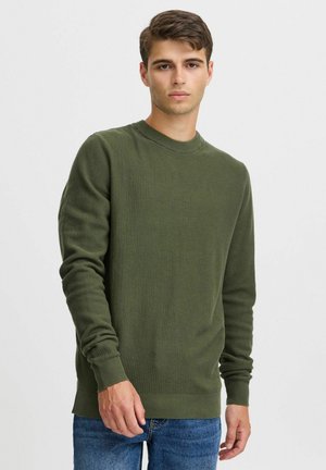 Olivengrüner, strukturierter Pullover mit rundem Halsausschnitt, gerippten Bündchen und Saum, kombiniert mit blauen Jeans, getragen von einem stehenden Modell.