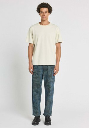 Homme debout face à l'avant portant un t-shirt beige clair uni, un pantalon camouflage bleu-vert et des chaussures noires sur un fond blanc.