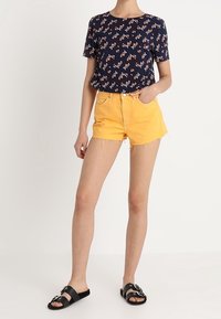 Námořnicky modrý květinový top s krátkým rukávem v kombinaci se žlutými denimovými šortkami s roztrhanými lemy a černými nazouvacími sandály se stříbrnými dekoracemi.