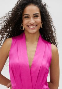 Vestido sin mangas en fucsia con un escote en V profundo, que presenta pliegues y una textura suave. Pendientes geométricos dorados acentúan el look.