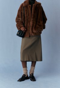 Hnědá bunda z umělé kožešiny, olivově zelená sukně, pletené krémové ponožky, černé mokasíny s texturovaným detailem a malá černá kabelka.