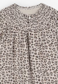 Blusa beige con estampado de leopardo negro y marrón, con cuello redondo fruncido y dos botones blancos en el cuello.