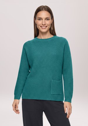 Jeune femme aux cheveux bruns raides portant un pull tricoté bleu sarcelle avec une poche avant et un pantalon sombre, souriant à la caméra.