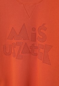 Orange pullover med en tekstureret overflade, der har hævede geometriske tekster i kontrasterende orange nuancer. Ribbet krave og V-formet udskæring.