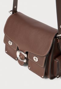 Brown leather shoulder bag with a flap closure, silver hardware, contrasting stitching, and side pockets. Compact design with a structured shape.