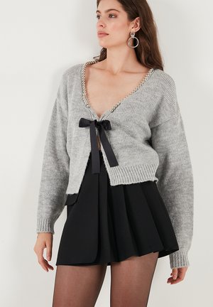 Gray knitted cardigan with a V-neck, embellished with pearl accents and a black ribbon tie at the front. Black pleated skirt underneath.