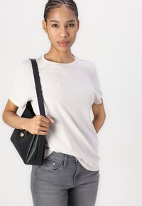 Camiseta de algodón gris claro con mangas cortas que presenta un sutil logo en relieve. Combinada con jeans grises y un bolso estructurado negro.