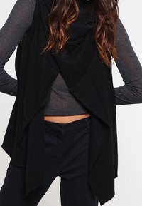 Svart stickad väst med öppen front och draperad design. Har en texturerad mönster. Buren över en grå långärmad topp och svarta byxor.