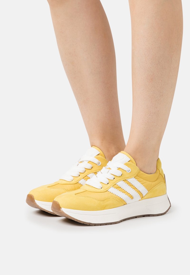 Personne portant des baskets de sport jaunes et blanches avec des semelles blanches épaisses sur un fond clair.