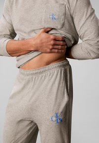 Personne soulevant un t-shirt gris à manches longues pour montrer l’abdomen, portant un ensemble gris assorti Calvin Klein avec logo sur la poche poitrine et le pantalon.