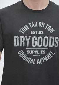 Άτομο που φοράει σκούρο γκρι μπλουζάκι με λευκό κείμενο που γράφει «Tom Tailor TRM Est. 62 Dry Goods Supplies Original Apparel.»