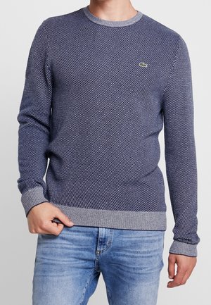 Mężczyzna ubrany w granatowy sweter z dzianiny o fakturze, z szarymi ściągaczami na mankietach i u dołu, w połączeniu z jasnoniebieskimi jeansami.