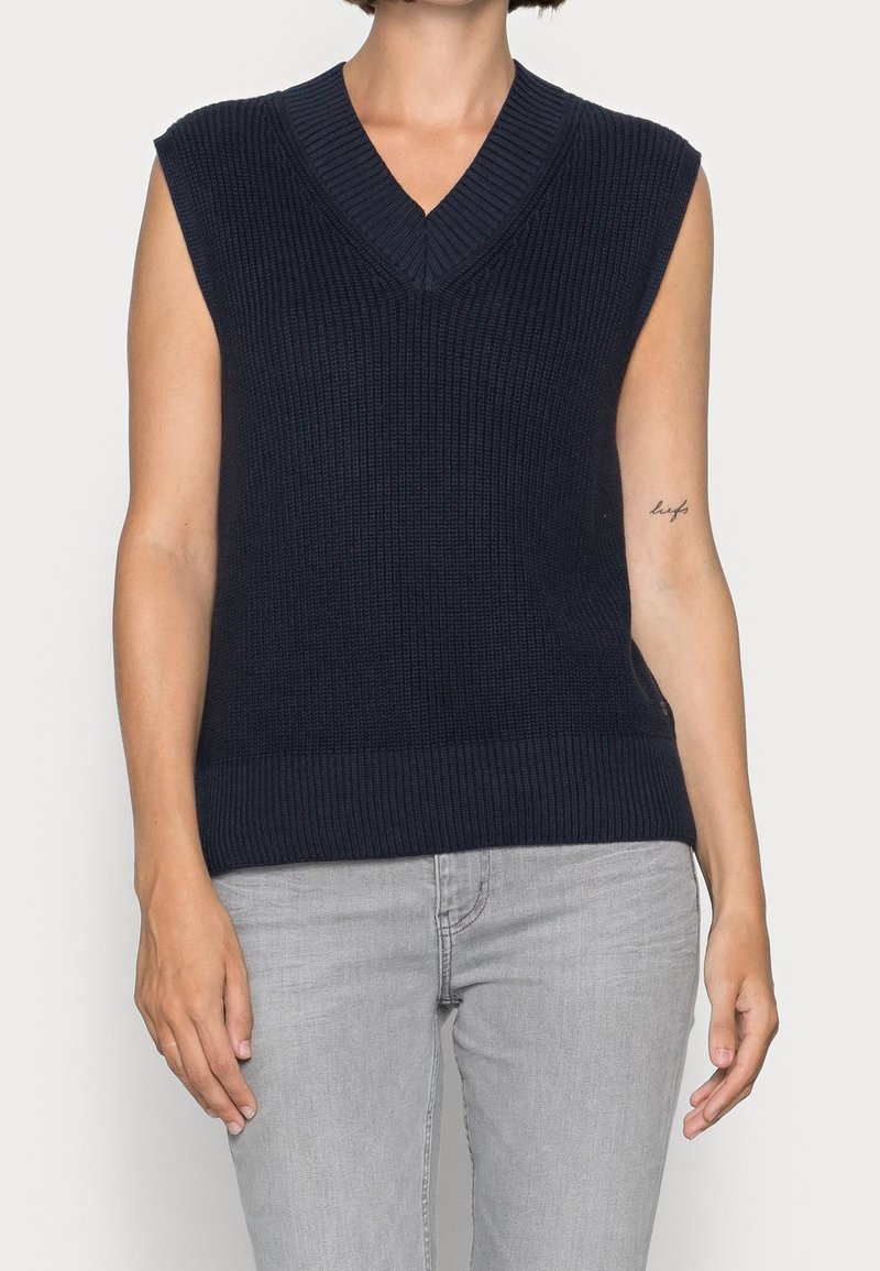Navy blue sleeveless sweater with a ribbed texture, V-neck design, and a fitted bottom hem. Worn over light gray pants.