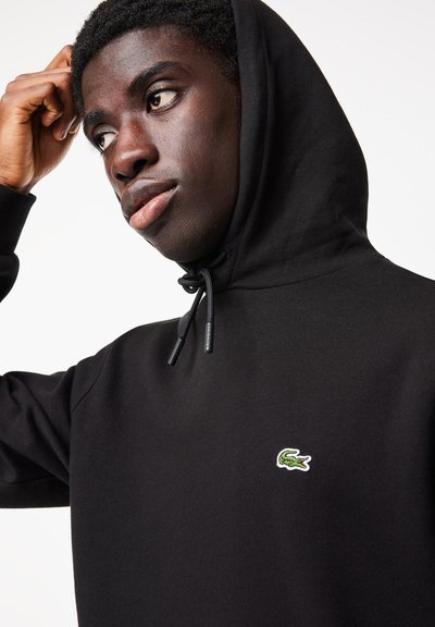 Hoodie noir avec cordon de serrage, arborant un petit logo de crocodile Lacoste vert et blanc sur la poitrine. Texture de tissu lisse.