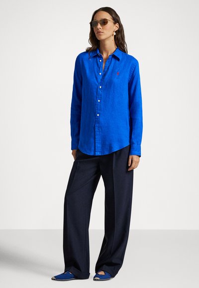 Μπλε λινού πουκάμισο με κουμπιά μπροστά, μυτερό γιακά και μακριά μανίκια, συνδυασμένο με σκούρο παντελόνι με φαρδιά πόδια και μπλε παπούτσια.