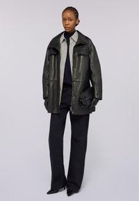 Giacca oversize in pelle nera con bottoni a pressione, camicia a righe sotto, pantaloni neri aderenti e una borsa nera strutturata.