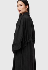 Svart oversizeklänning med rynkad rygg, långa ärmar och krage. Mjuk textur med subtil lyster. Har en sidoficka och minimalistisk design.