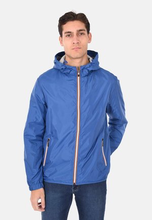 Giovane uomo con giacca a vento con cappuccio blu con cerniere e tasche rifinite in arancione, in piedi con una mano dietro la schiena.