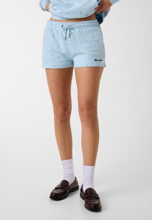Person trägt hellblaue Shorts mit Blumenmuster und Kordelzug, weiße Socken und dunkelbraune Loafers, steht vor einem einfarbigen Hintergrund.